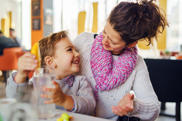 Obraz na płótnie Canvas Young woman mother with small boy child son having fun at cafe or restaurant caucasian kid smiling while sitting by the table looking at his mother while talking