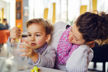 Obraz premium Portrait of small three years caucasian boy kid child sitting by the table with his mother young woman female watching at cafe having a glass of water son draining lemon making lemonade