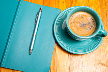 Top down view of a turquoise cup and saucer filled with coffee next to a turquoise pen and notebook on a wooden table top