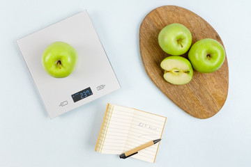 Fresh green apple on gray digital kitchen scales. Near notepad with number of calories, pen and some apples on wooden board. Flat lay. Kitchen equipment. Healthy food concept. Weighing products. - Powered by Adobe