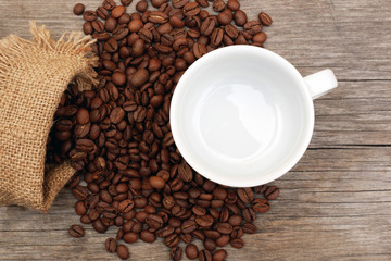 top view on coffee beans spilled from burlap sack and white empty cup on aged wooden background