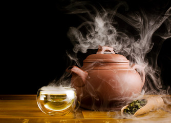 Chinese tea ceremony. Clay teapot with green oolong tea Tieguanyin, scoop of tea, glass cup of hot tea on a black background with vapour.