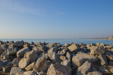 Many large stones on the seashore against the background of water and the city in the distance.