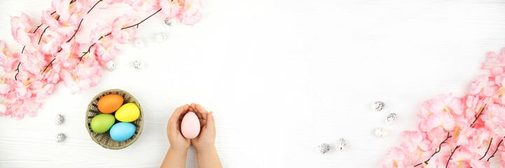 Banner made from pastel colored Easter eggs in the nest with the branch of pink flowers on white wooden background. Child's hands holding pink egg on the Easter background.