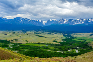 Fototapeta premium Panoramic view of mountain range under cloudy sky. Plateau with green trees and small rivers against snow-capped peaks.