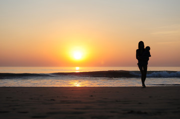 Fototapeta premium Silhouette of woman with a child in her arms on the background of sunset by the sea. Young slender girl carries a baby in the rays of setting sun. Beautiful mother walking along the sandy beach.