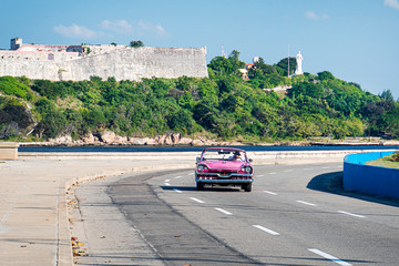 Pink old cabriolet rides along Malecon embankment against the background of the old Fort. el Morro lighhouse