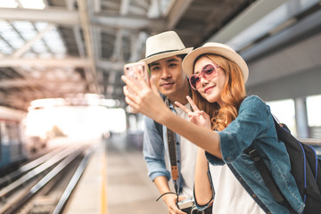 Happy Asian couple traveler holding a mobile phone in station and waiting for train in vacation...