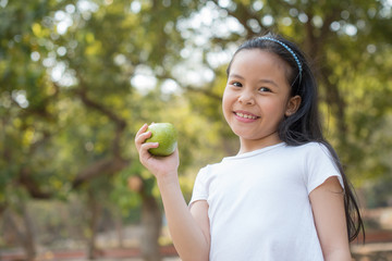 photo happy Little asian girl child standing with big smile. holding green apple in your hand.fresh healthy green bio background with abstract blurred foliage and bright summer sunlight