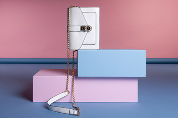 A small women's rectangular handbag on a chain strap stands on a pink, blue stand. Studio photo