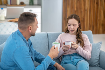 Daughter talking to father sitting on couch looking into cup.