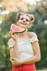 Fototapeta premium A stylish beautiful young woman with bright juicy makeup in the form of a watermelon holds a piece of watermelon in her hands