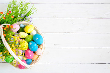 Colorful easter eggs in basket and flowers on white wooden table. Top view with copy space