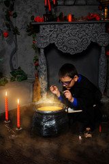 A wizard boy in glasses with a magic wand in his hands and in a black mantle with a hood is cooking a potion in a black cauldron, sitting in fog and smoke.