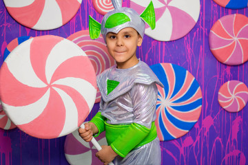 A boy in an alien costume holds a big candy in his hands against the background of a wall of sweets.