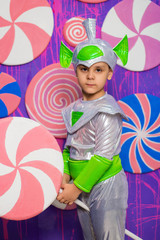 A boy in an alien costume holds a big candy in his hands against the background of a wall of sweets.