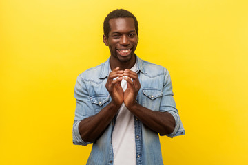 Portrait of sneaky devious man in denim casual shirt with rolled up sleeves smiling, twiddling fingers and thinking cunning idea, planning evil trick. indoor studio shot isolated on yellow background