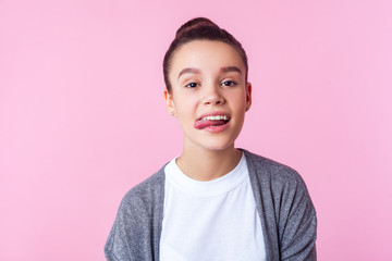 Portrait of carefree childish brunette girl with bun hairstyle in casual clothes showing tongue, disobedient teenager making face, having fun at camera. indoor studio shot isolated on pink background