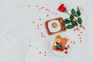 Pancakes in the shape of a heart with berries, morning  coffee and rose on white bed.  top view. Valentine's day breakfast concept