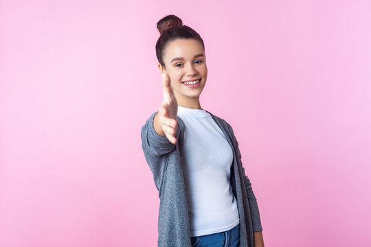 Welcome! Portrait of lovely brunette teenage girl with bun hairstyle in casual clothes giving hand to handshake, meeting new people with sincere toothy smile. studio shot isolated on pink background