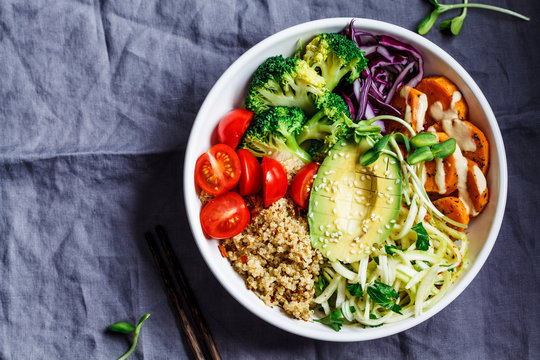 Buddha bowl salad with quinoa, avocado, broccoli, sweet potato and tahini dressing, gray background.