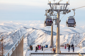 A gondola cableway with a two booths suspended on a cable with people on the observation deck and snow on winter. Manzherok Ski resorts and snowboarding in Altai mountains. © Aleksandr Kondratov