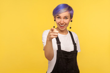 Hey you! Portrait of cheerful hipster woman with violet short hair in denim overalls pointing finger at camera and smiling, making choice, selecting you. isolated on yellow background, studio shot