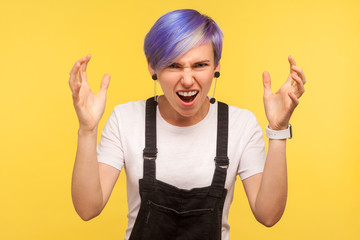 Portrait of aggressive hipster girl with violet dyed short hair in denim overalls quarreling with raised hands, screaming in anger with mad and furious face. isolated on yellow background, studio shot © khosrork