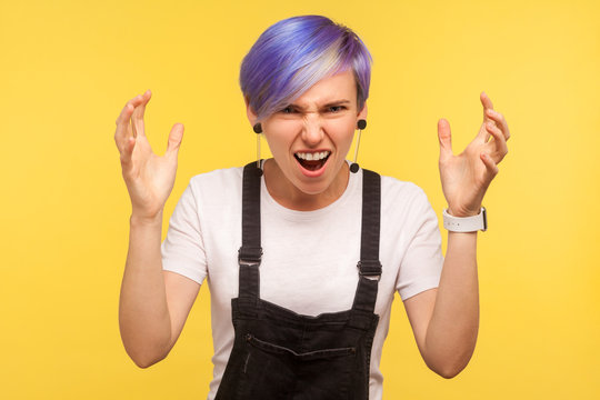 Portrait of aggressive hipster girl with violet dyed short hair in denim overalls quarreling with raised hands, screaming in anger with mad and furious face. isolated on yellow background, studio shot