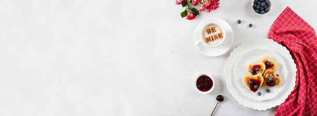 Valentine pancakes in heart shape with jam and berries on white plate with a cup of coffee and roses bouquet. St. Valentines Day surprise, breakfast concept. Cafe menu banner. Copy space