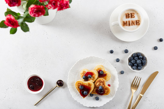 Valentine's day breakfast or brunch. Homemade heart shape pancakes with cherry jam and blueberries a cup of coffee and bouquet of roses. Top view, copy space. Cafe, restaurant menu, recipe