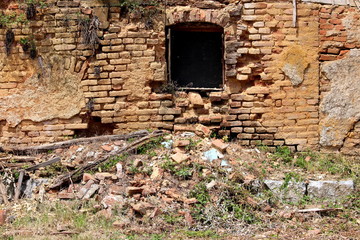Fototapeta na wymiar Broken window mounted on red brick wall of ruins of destroyed old wooden and bricks suburban family house mixed with small plants in old part of town