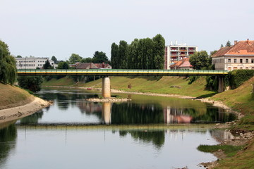 Fototapeta na wymiar Green and yellow old metal bridge mounted on strong concrete foundation over local river with low summer water levels surrounded with apartment buildings and family houses among tall trees