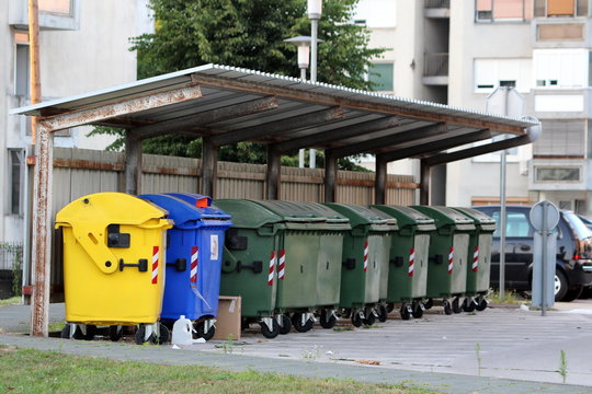 Row of various large plastic trash containers for ecologically sorting garbage for recycling under metal roof tiles protection between old tall apartment buildings