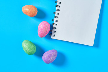 Many scattered colorful eggs with glitters near blank paper notepad for message lies on blue desk on kitchen. Easter concept. Space for text. Top view
