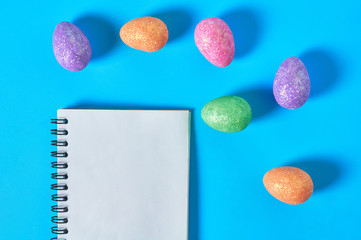 Many scattered colorful eggs with glitters near blank paper notepad for message lies on blue desk on kitchen. Easter concept. Space for text. Top view