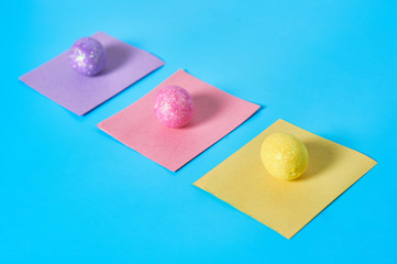 Row of three colorful eggs with glitters and square paper stickers lies on blue desk on kitchen. Easter concept. Close-up