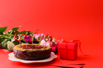 Concept Women's Day or St. Valentines Day. Fresh roses and gift box on a red background and a chocolate cake with candles 