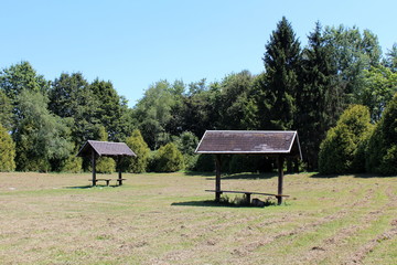 Two dilapidated wooden public benches with makeshift roofs covered with broken roof tiles at local public park surrounded with freshly cut grass and dense trees