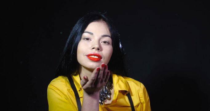 Beautiful brunette with brown eyes, long black hair, red lipstick and manicure in yellow blouse blowing loose glitter sparkles from her hand that fly into camera forming a brilliant cloud slow motion