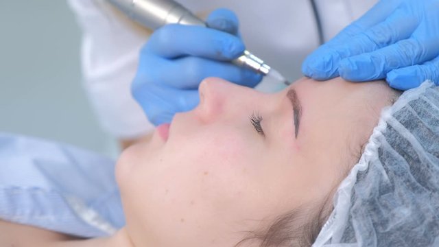 Cosmetologist making eyebrows microblading procedure in beauty salon for girl using tattoo apparatus. Beautician in gloves is doing permanent eyebrow makeup for woman, side view. Beauty industry.