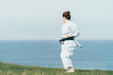 Back view of unknown young female redhead karate athlete training on top of cliff with sea on...