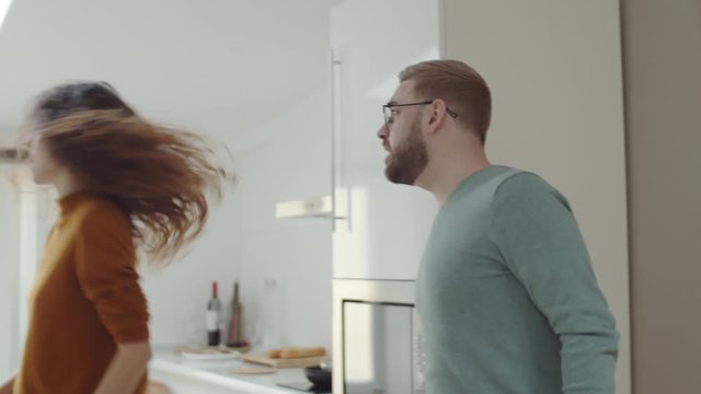 Waist-up tracking shot of married Caucasian couple having explosive fight in kitchen at home, angry man shouting at wife and furious woman screaming then picking up glass of water and drinking