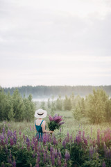 woman in a hat in a field with a bouquet of lupins in the morning in the fog.