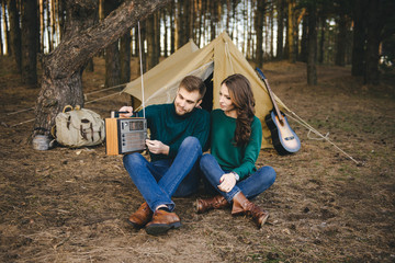 Young couple in love camping tourists sitting by a fire against a tent in the forest listening to retro radio