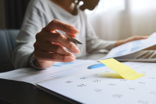 Asian woman will be write on yellow paper notes and put a warning on calendar. The other hand hold the report graph and has laptop on the table for preparing data to presentation marketing in meeting