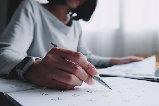 Asian woman will be write on calendar paper note and put a warning on work sheet. Other hand hold the report graph and has laptop on the table for preparing data to presentation marketing in meeting