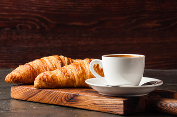 Espresso in a white cup and two croissants on dark background, on a wooden board, place for text, side view