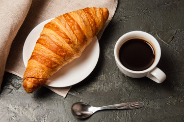 Croissant and cup of coffee with spoon and saucer on a dark background with gray linen cloth