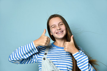 Teenage female in jeans overall, striped sweatshirt. She smiling, showing thumbs up, winking, posing on blue background. Close up